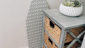 A corner of a room with a gray ironing board, a white plant pot, and a woven basket under a table.