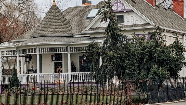 A two-story house with a wrap-around porch, a chimney, and a large evergreen tree in front.