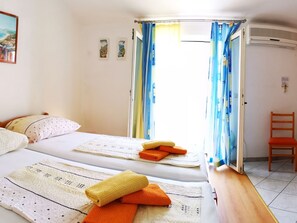 Ein Zimmer mit zwei Betten, einem Holzstuhl und Blick ins Freie durch ein Fenster mit Vorhängen.
