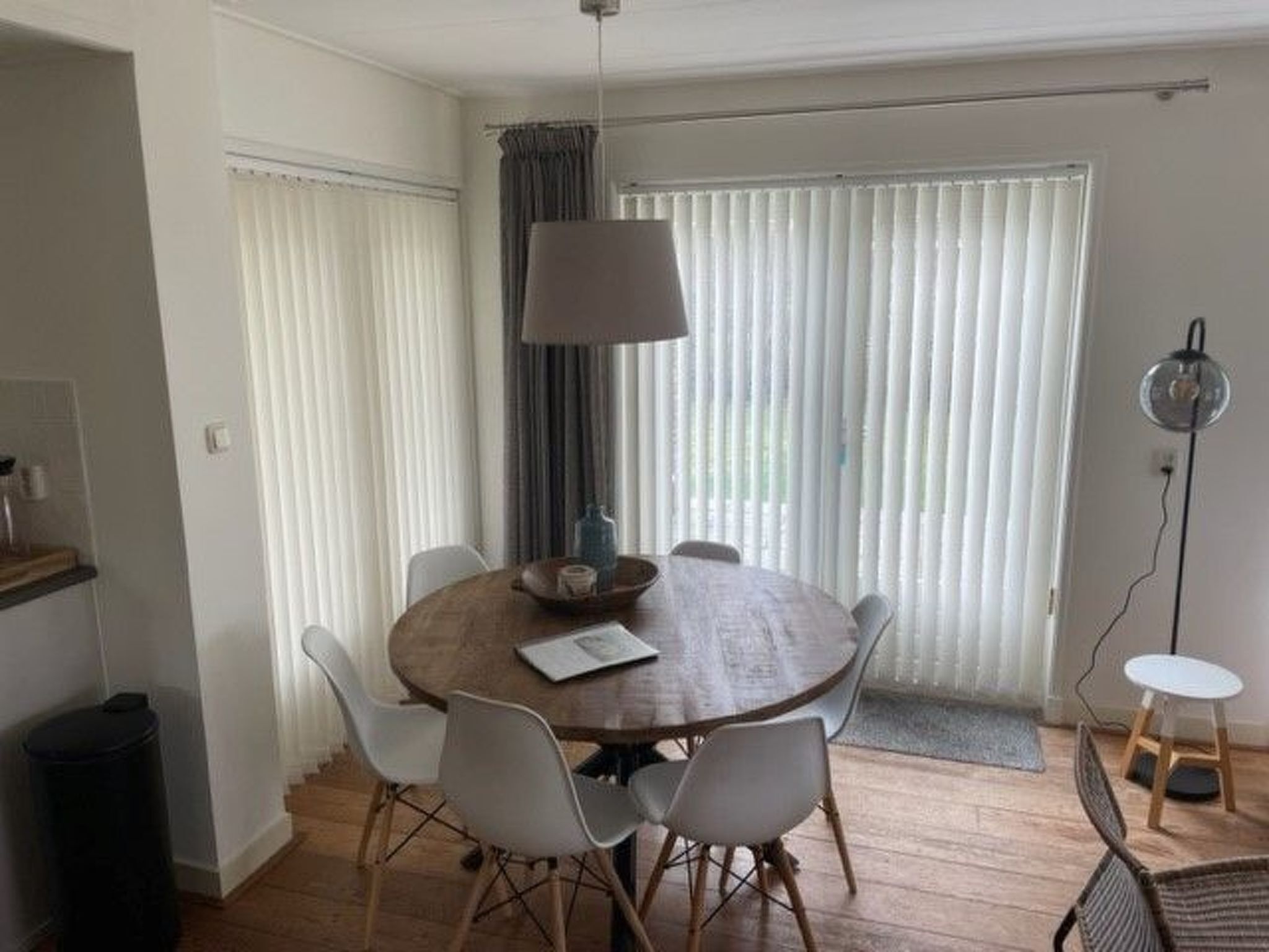 Spiseområde med et rundt træbord, seks hvide stole, en lampe og et vindue med persienner.
