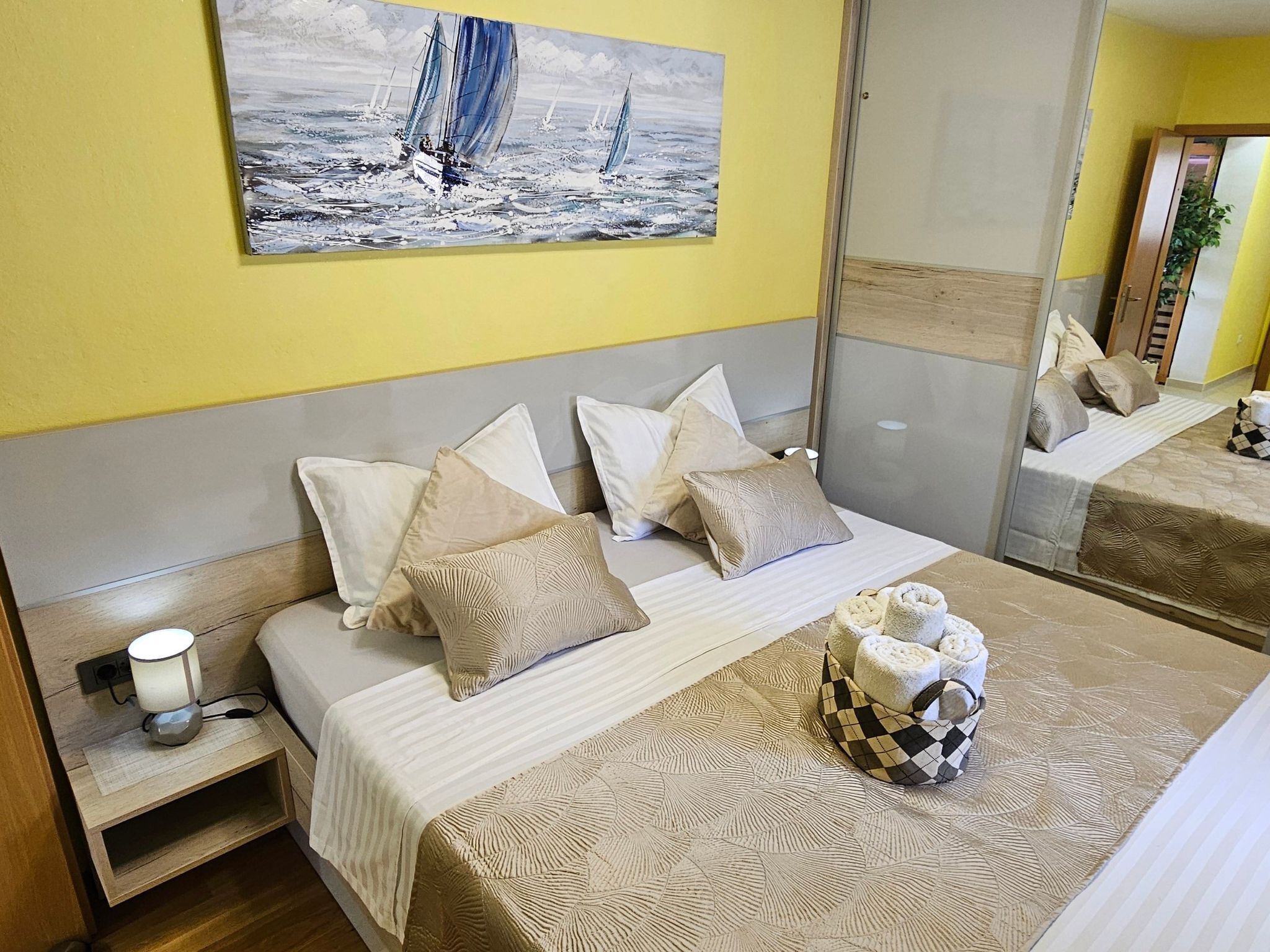 Ein Schlafzimmer mit Bett, Kissen, einem Nachttisch mit Lampe und einem Bild von Segelbooten an der Wand.