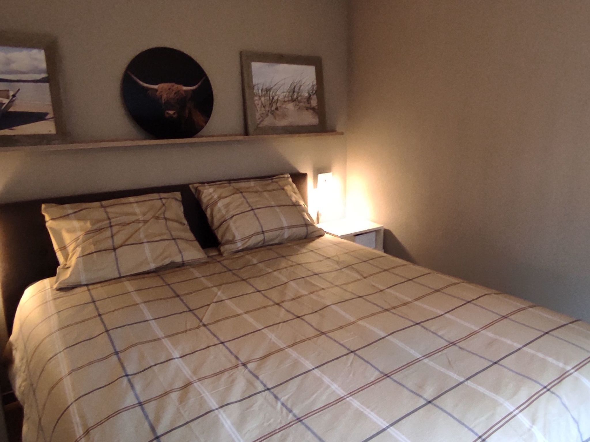 Un lit bien fait, recouvert d’une couverture à carreaux, une lampe de chevet et deux tableaux encadrés accrochés au mur au-dessus du lit.