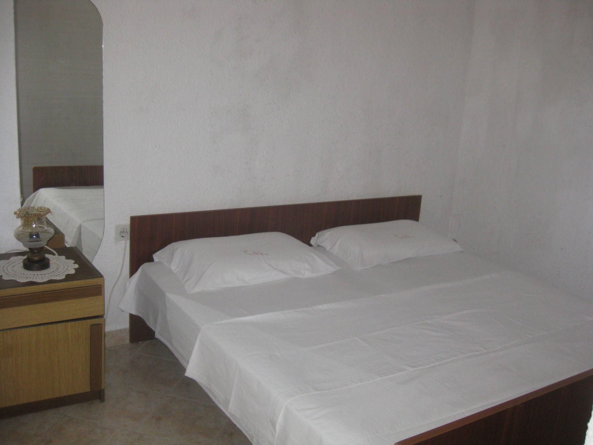 Een eenvoudige, nette kamer met een enkel bed, een houten nachtkastje en een bijzettafeltje met een lamp.