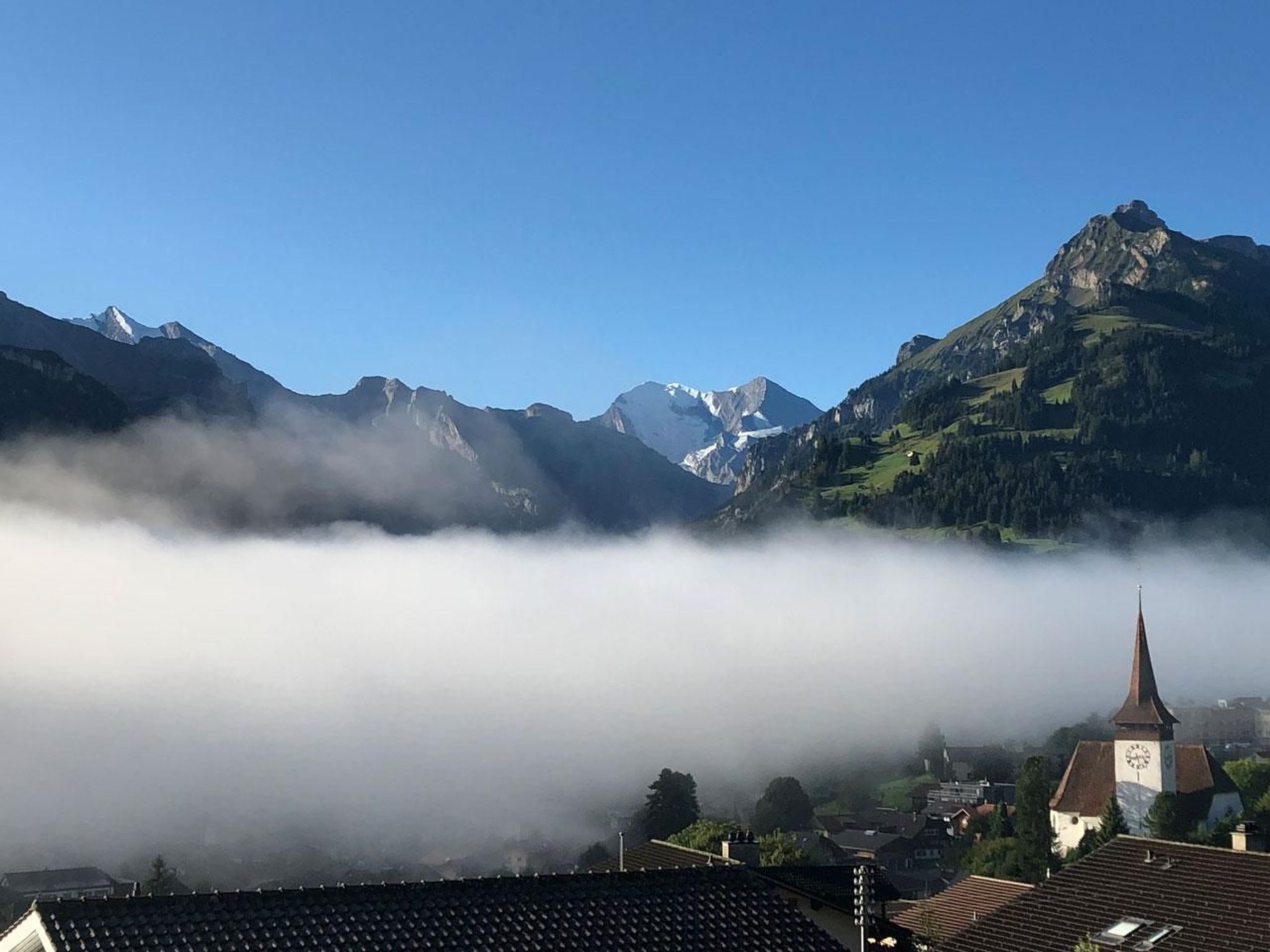 Un paysage de montagne avec une église et des bâtiments au premier plan, entourés de nuages et de sommets enneigés.