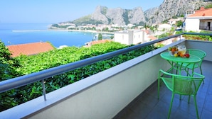 Un balcon avec des chaises vertes et une table, d’où l’on peut admirer une ville côtière aux eaux bleues et limpides, avec des montagnes rocheuses en arrière-plan.