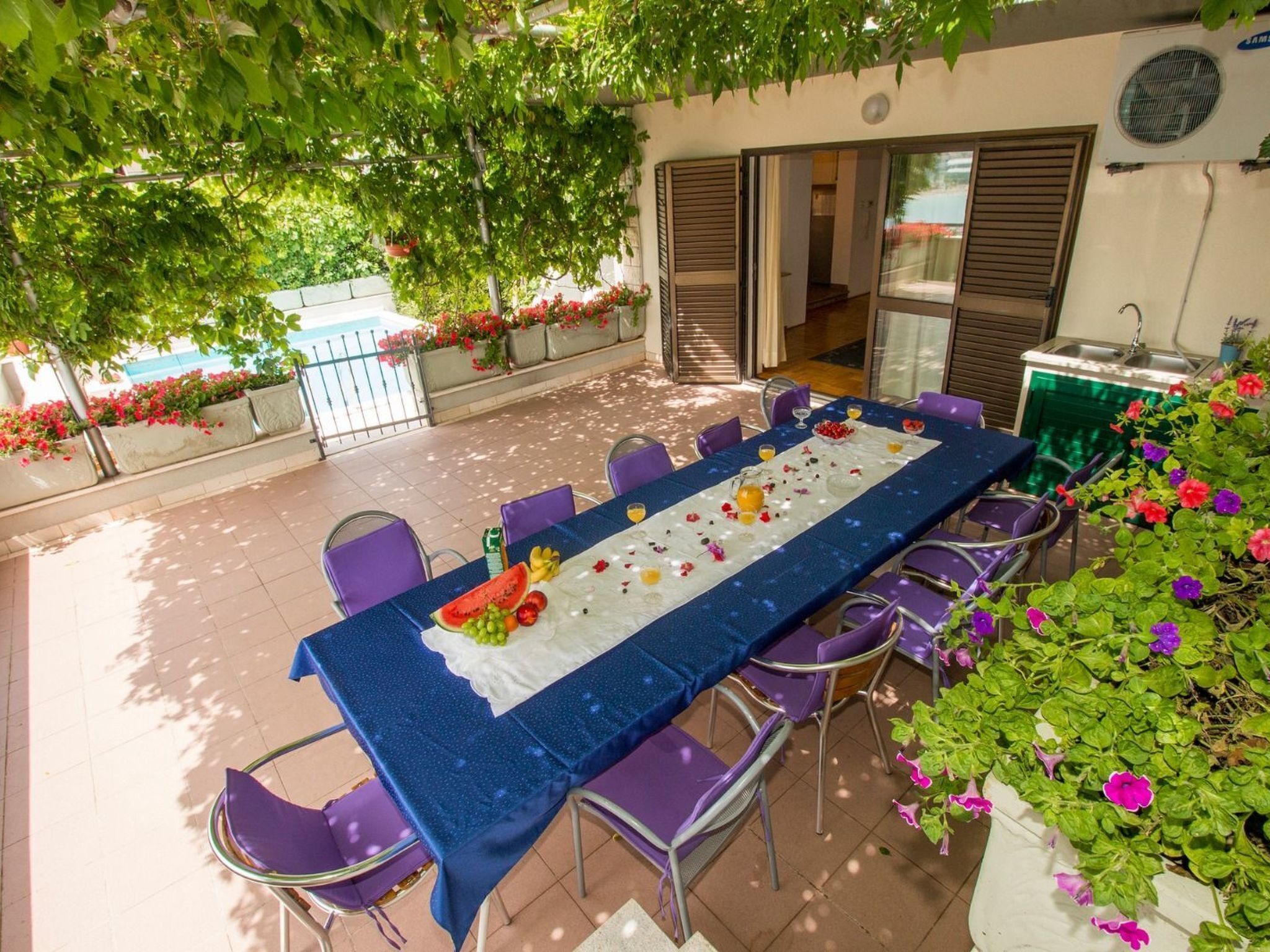 Un espace de restauration en plein air avec une longue table dressée pour un repas, entourée de chaises et décorée de fleurs et de fruits frais.
