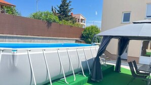 Una piscina elevada con estructura blanca y revestimiento azul, rodeada de césped artificial verde, con una mesa y sillas de jardín cerca.