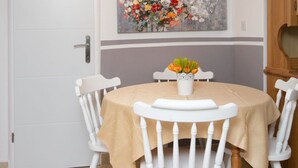 Un coin repas avec une table ronde, des chaises blanches et une nappe beige. Un tableau représentant des fleurs est accroché au mur, au-dessus de la table.