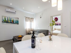Ein modernes Wohnzimmer mit einem weißen Sofa, einem Esstisch mit einer Weinflasche und Gläsern, einem Gemälde von Segelbooten und einer Blumenvase.