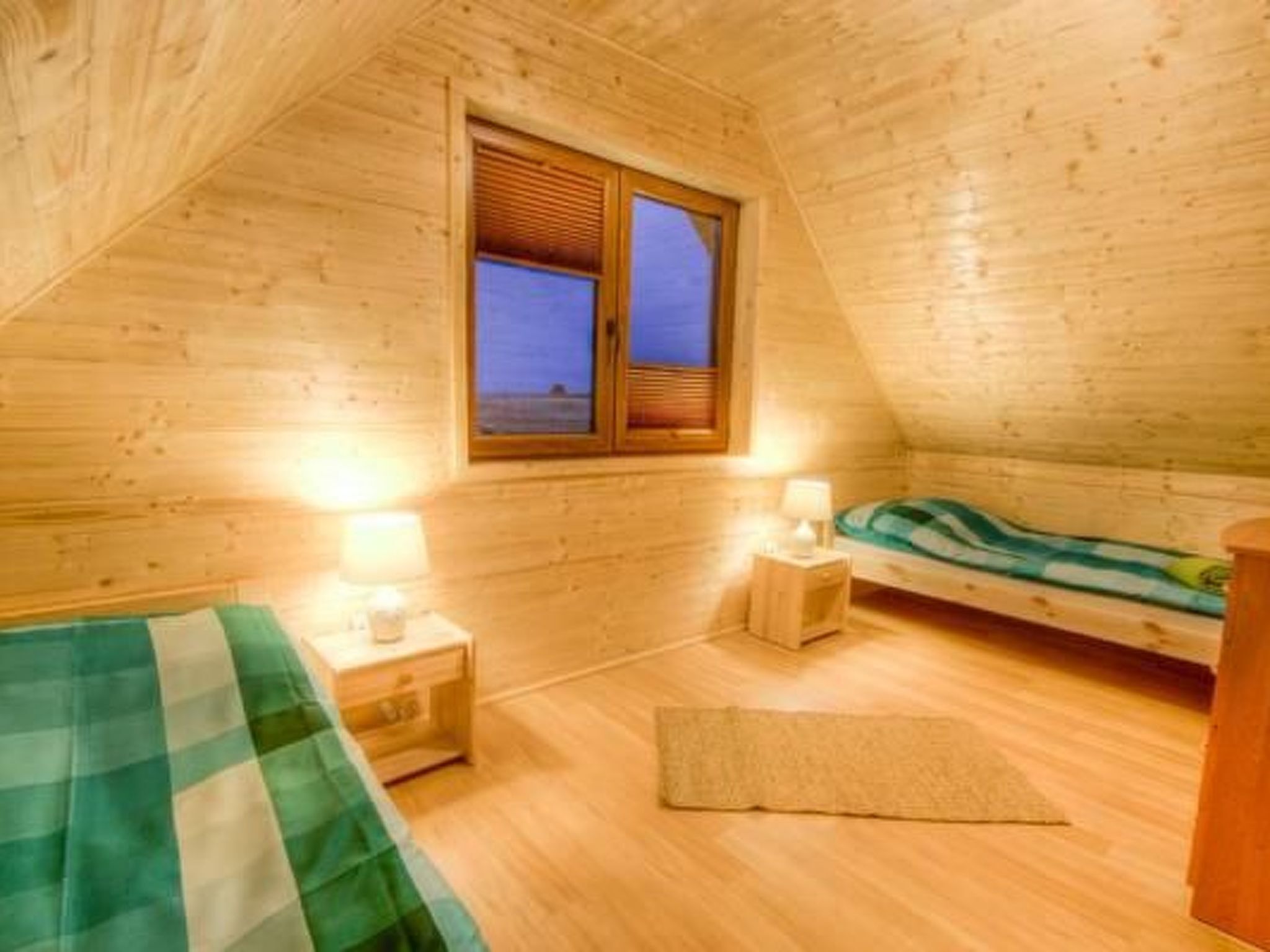 A cozy, single-room cabin with a sloped ceiling, a window with shutters, a bed with a green and white checkered blanket, a bedside table with a lamp, and a small rug on the wooden floor.