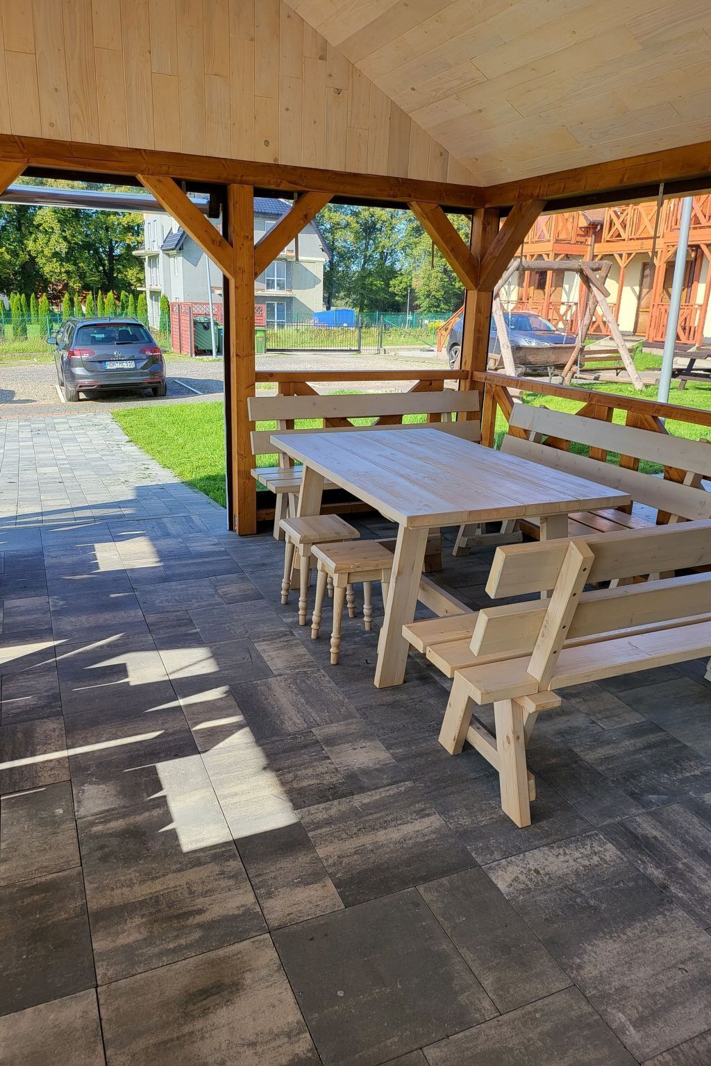 Ein überdachter Picknickbereich mit Holzbänken und -tischen, einem gefliesten Boden und Blick auf eine Straße mit parkenden Autos und Gebäuden im Hintergrund.