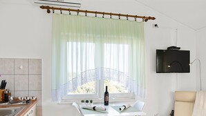 Une cuisine moderne avec un coin repas, un téléviseur fixé au mur et une vue sur l’extérieur grâce à une fenêtre ornée de rideaux légers.