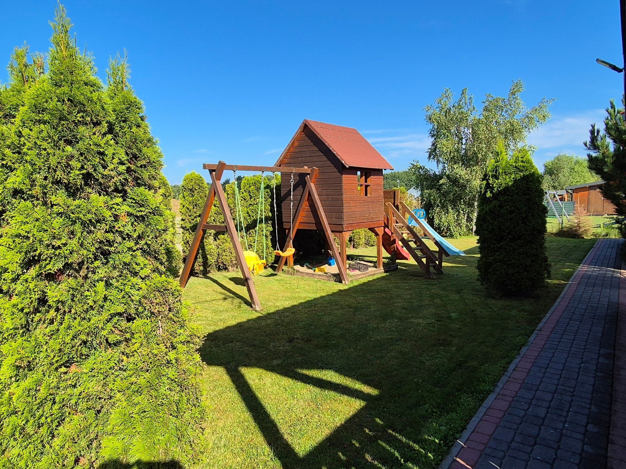 Una casita de juegos de madera con techo rojo y un tobogán, rodeada de un césped cuidado y árboles.