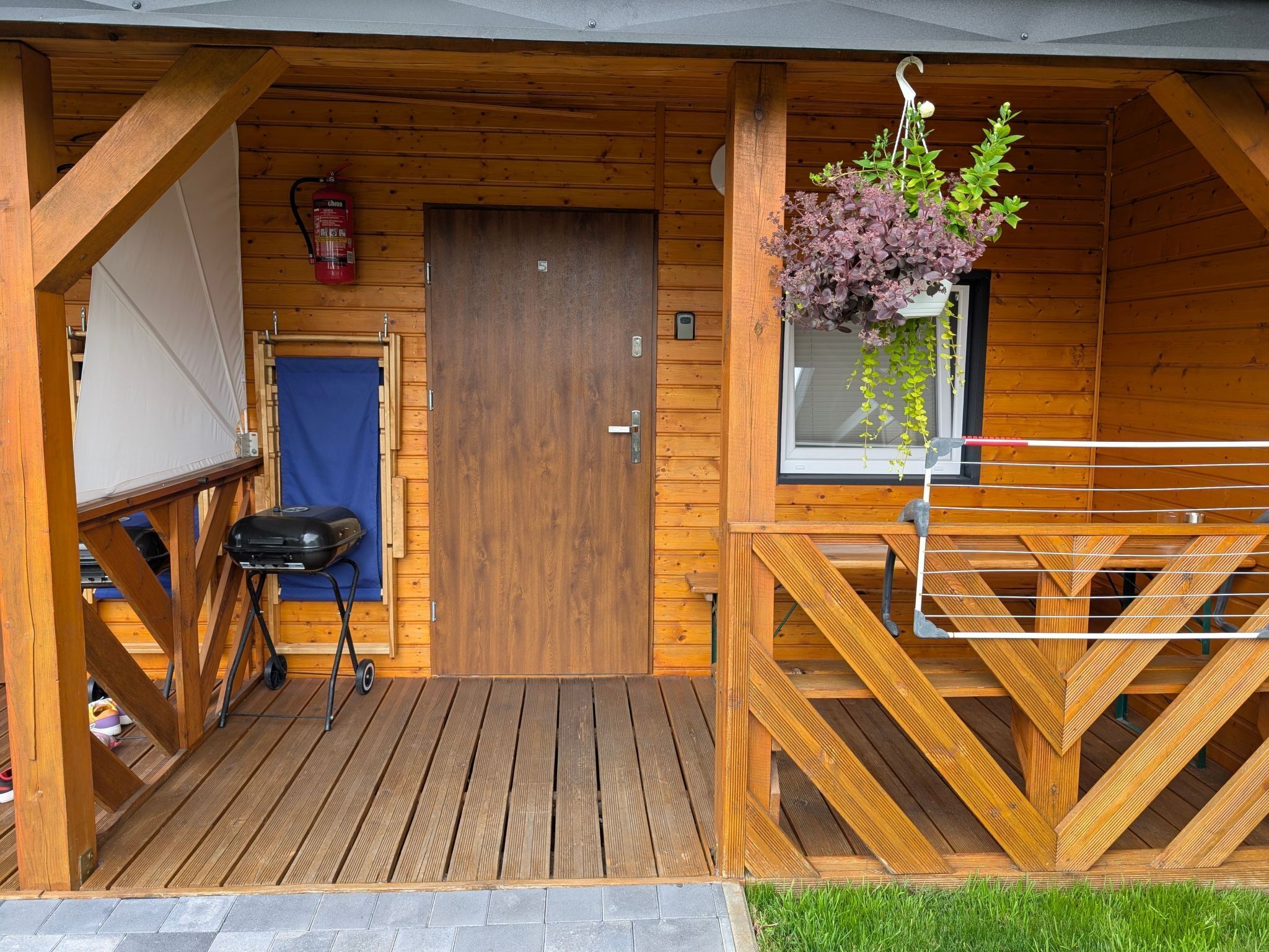 Eine Holzhütte mit Tür, einer kleinen Veranda, einem Grill und einer hängenden Pflanze.