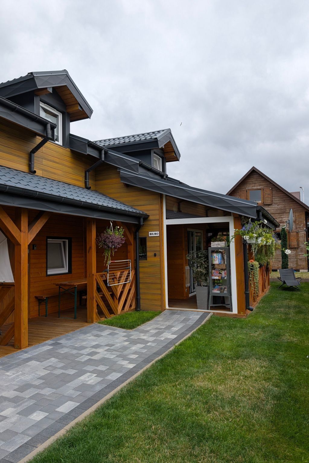 Un edificio de madera con un porche cubierto, un pequeño espacio de estar al aire libre y un sendero pavimentado que conduce a la entrada.