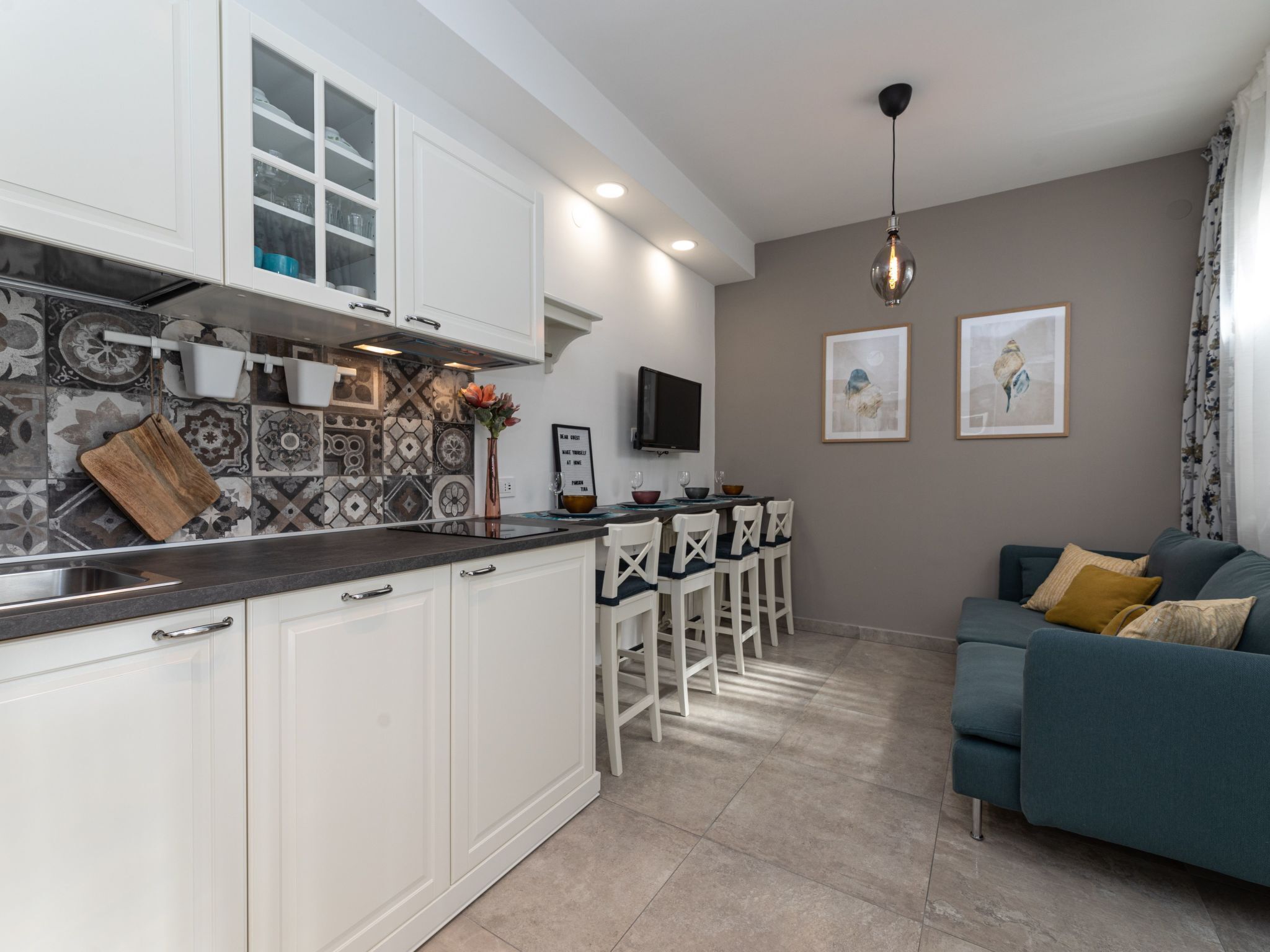 Una cocina moderna con armarios blancos, encimera negra y salpicadero estampado. Hay una zona de comedor con sillas blancas y una barra, además de una sala de estar con un sofá azul y un cuadro en la pared.