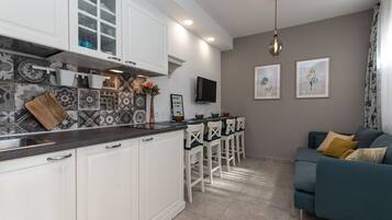 Una cocina moderna con armarios blancos, encimera negra y salpicadero estampado. Hay una zona de comedor con sillas blancas y una barra, además de una sala de estar con un sofá azul y un cuadro en la pared.