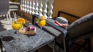 Ein Balkon mit zwei Tischen, jeder Tisch mit einem Getränk und einem Buch. Auf einem Tisch liegt ein Hut, auf dem anderen eine Sonnenbrille. Der Balkon hat eine weiße Brüstung und bietet Blick auf Berge im Hintergrund.