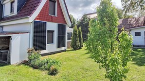 Ett tvåvåningshus med ett rött tak, vita väggar och en liten trädgård framför huset, med ett träd och några buskar.