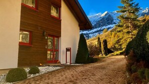 A modern two-story building with a wooden facade and red-framed windows, a paved driveway, and a mountainous backdrop. - Chalet Kristall by Interhome (Albinen)