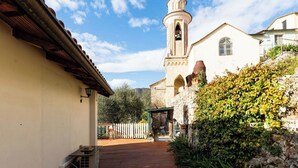 Un estrecho callejón que conduce a una iglesia con un campanario, rodeado de vegetación y muros de piedra.