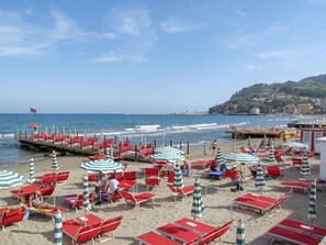 Ein Strand mit roten und weißen Streifen-Schirmen und Liegestühlen, eine Mole mit roten Sesseln sowie ein Blick auf das Meer und Gebäude im Hintergrund.