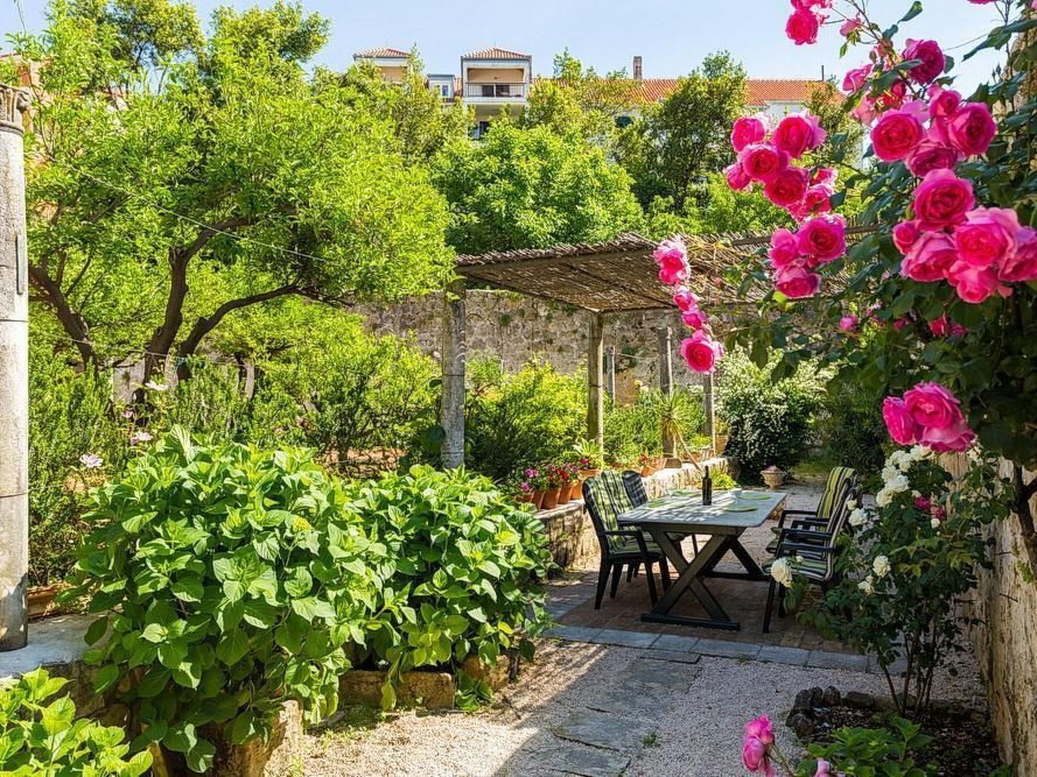 Un patio jardín con mesa y sillas, rodeado de abundante follaje y plantas en flor.
