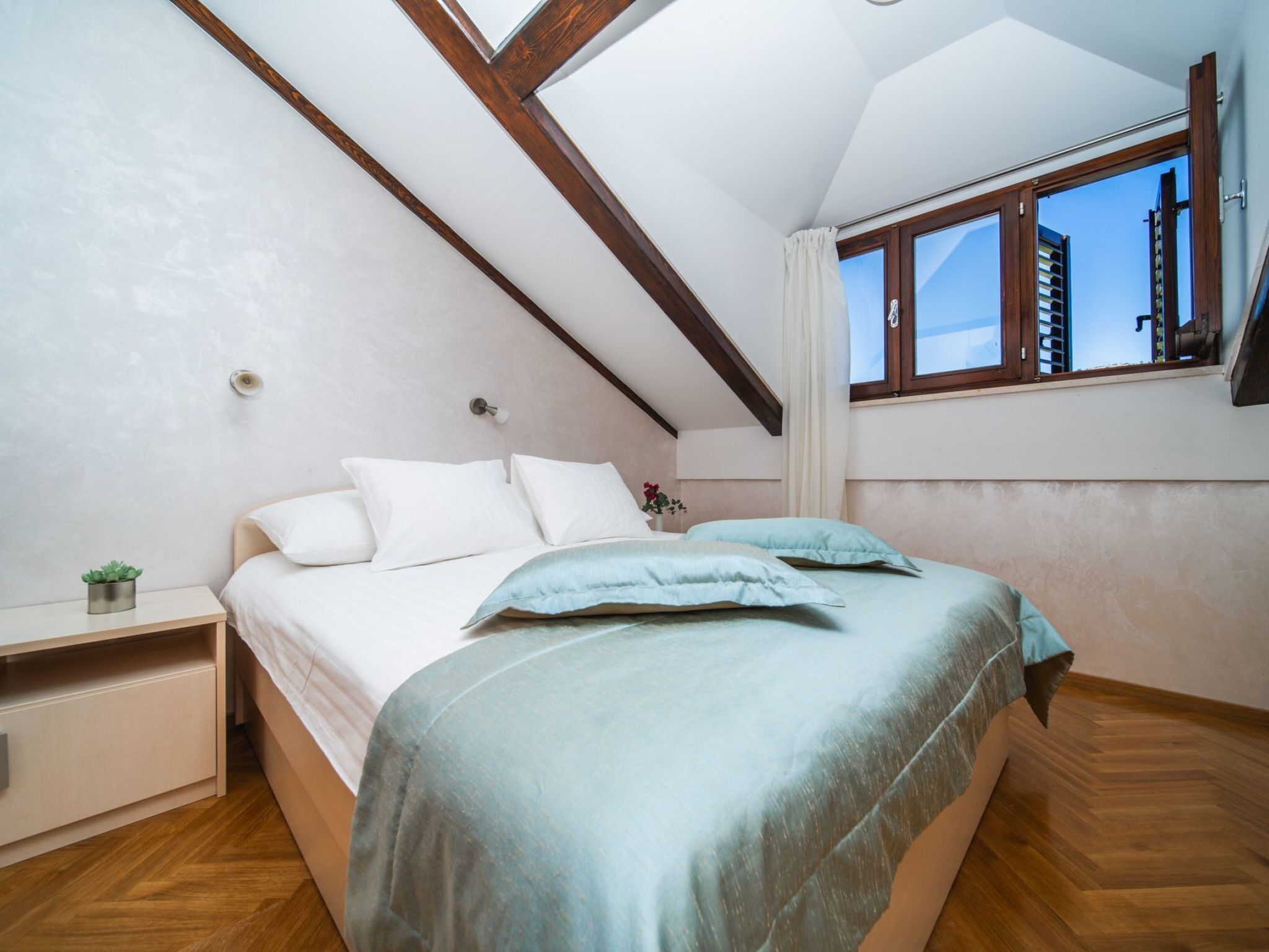 A bedroom with a sloped ceiling, a bed with white and teal bedding, a nightstand with a plant, and a window with a view of buildings.