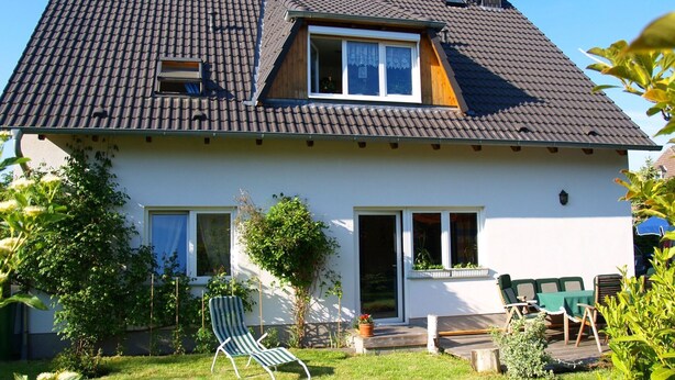 Ein zweistöckiges Haus mit geneigtem Dach, einer Veranda mit Tisch und Stühlen und einem Rasen mit einem einzelnen Liegestuhl.