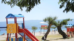 Un terrain de jeux équipé d’un toboggan et de structures d’escalade, avec en toile de fond une plage et des montagnes.