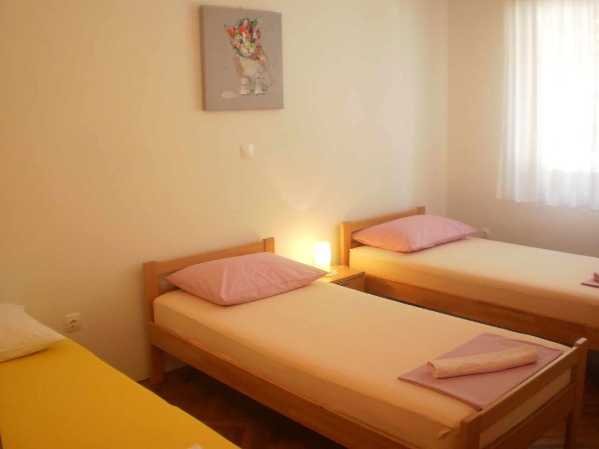 A room with two single beds, each with a wooden headboard and a pink pillow. There is a small bedside table with a lamp on one bed. A painting of a kitten hangs on the wall above the beds.