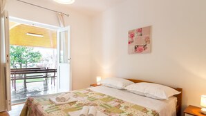 A bedroom with a bed, bedside tables, a wall-mounted artwork, and an open door leading to an outdoor area with a bench and a tree. - Carpe Diem by Interhome (Trogir)