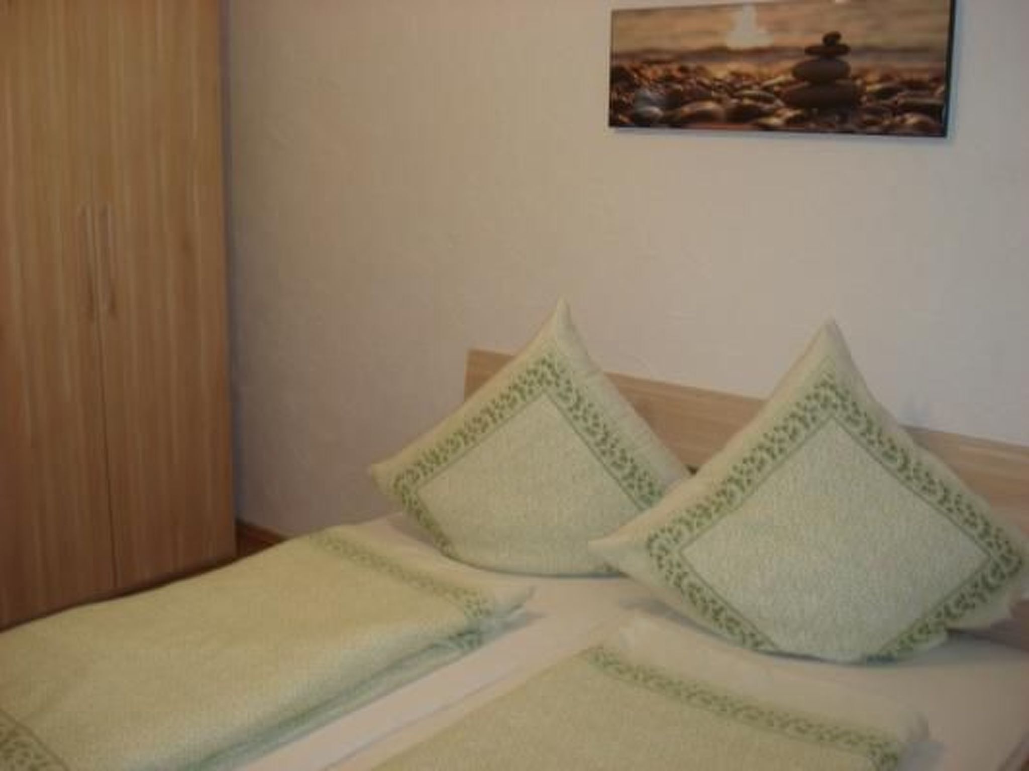 A bed with a green blanket and pillows, a wooden wardrobe, and a painting of a rocky landscape on the wall.