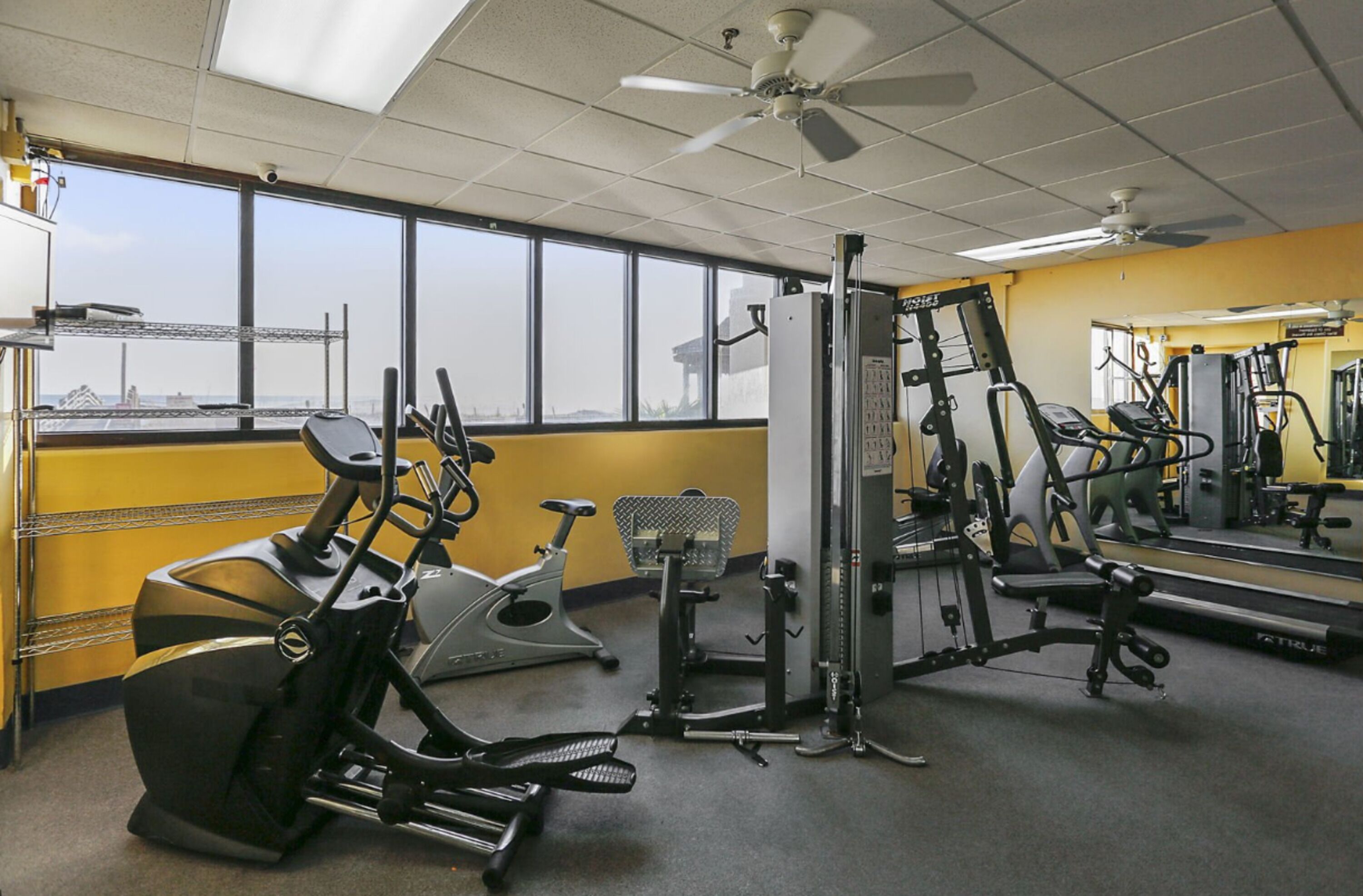 Ein Fitnessraum mit verschiedenen Trainingsgeräten, großen Fenstern und einem Deckenventilator.