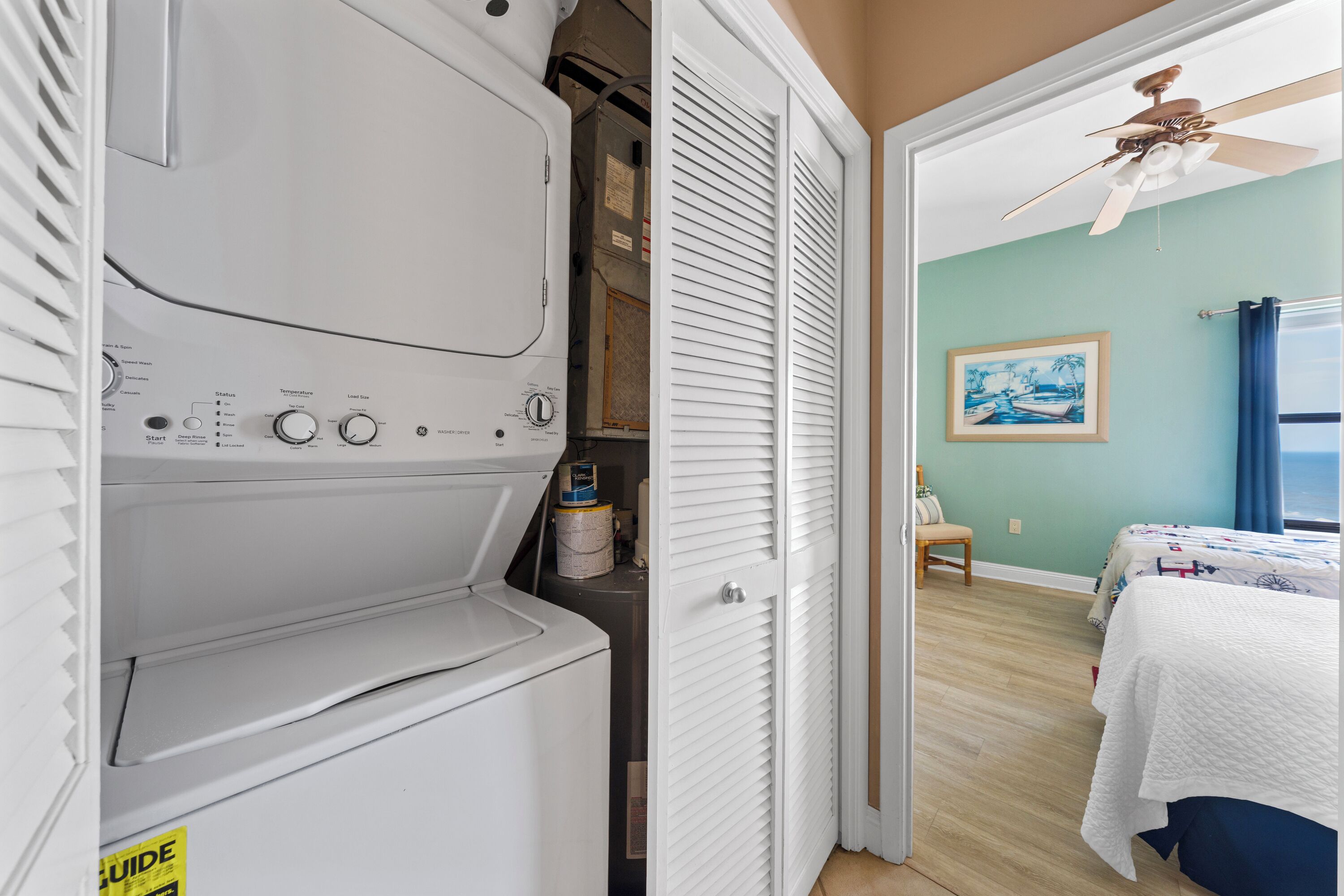 Une buanderie équipée d’un lave-linge et d’un sèche-linge, un placard avec une porte blanche, et une chambre à coucher comprenant un lit, une chaise et un ventilateur de plafond.