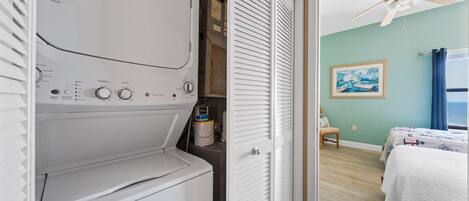 Une buanderie équipée d’un lave-linge et d’un sèche-linge, un placard avec une porte blanche, et une chambre à coucher comprenant un lit, une chaise et un ventilateur de plafond.