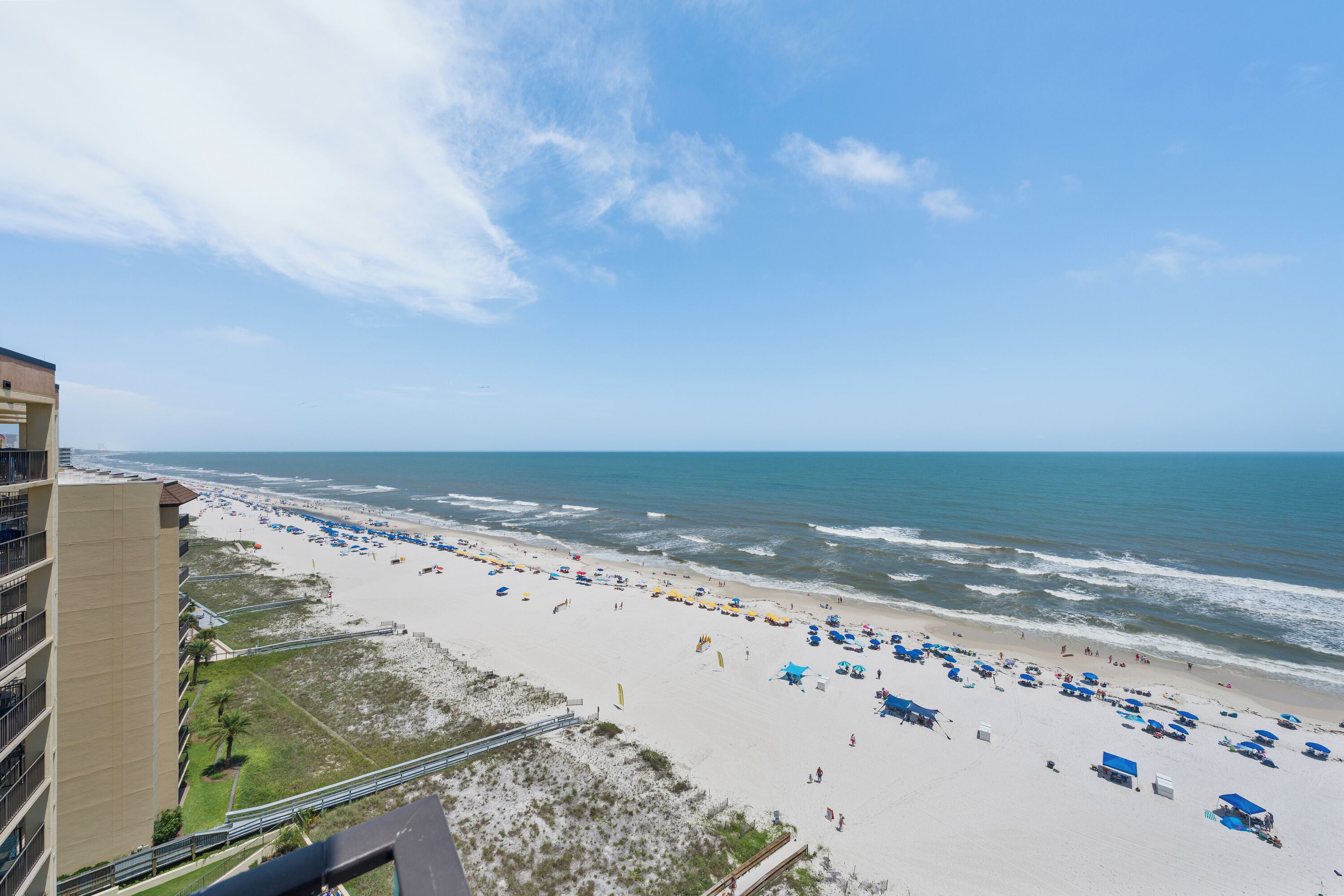Une plage avec des parasols et des gens, un immeuble avec des balcons, et un ciel dégagé.