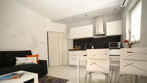 Una sala de estar moderna con un sofá negro, una mesa de centro blanca y juguetes coloridos sobre una alfombra. Se ve un área de cocina con armarios blancos, una encimera negra y electrodomésticos de acero inoxidable.