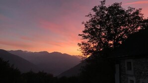 Um edifício de pedra com telhado inclinado, emoldurado por uma paisagem montanhosa durante o pôr do sol.
