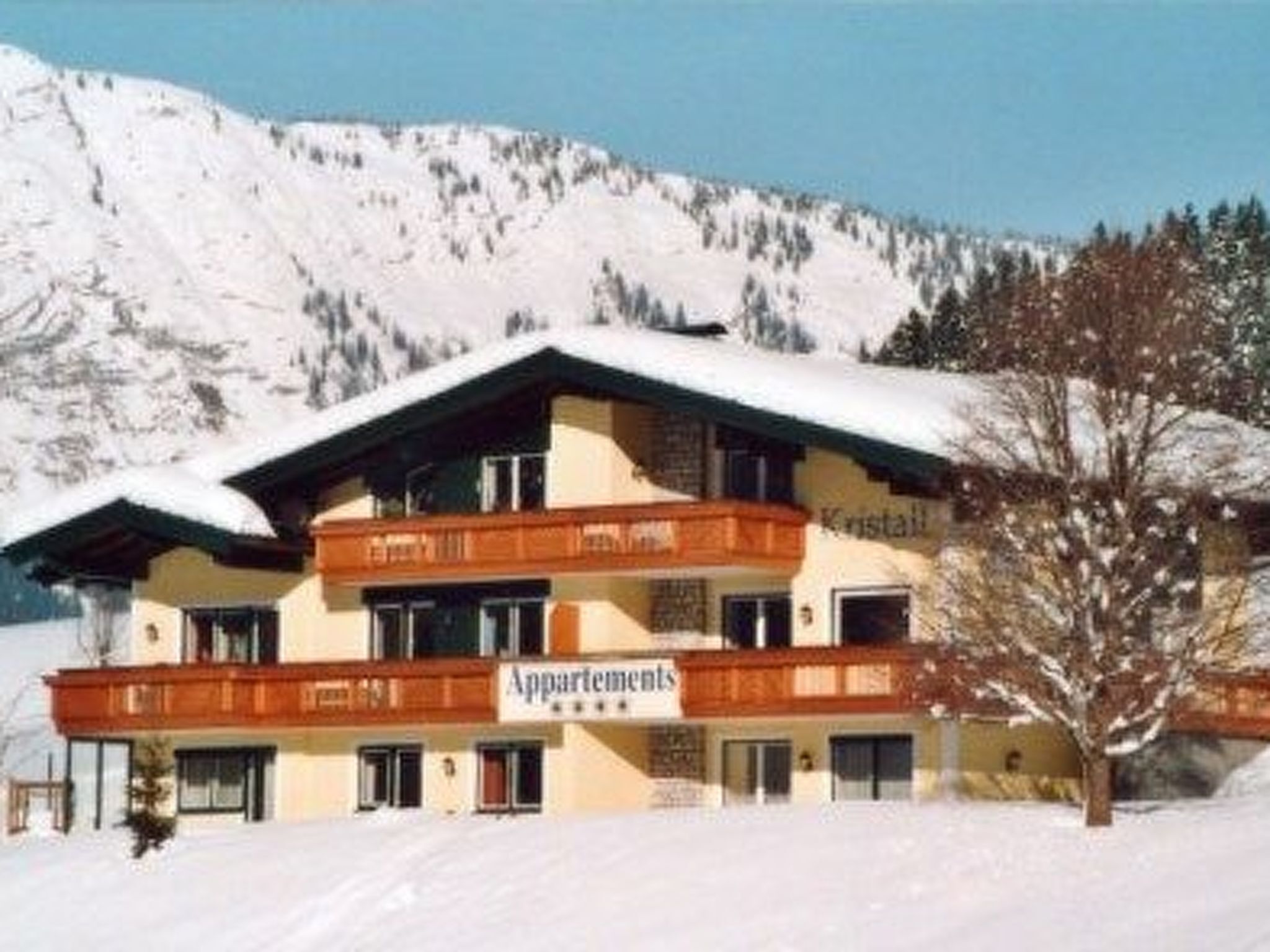 Ein mehrgeschossiges Gebäude mit einem Schild, auf dem "Apartments" und "Kristall" steht. Es hat ein geneigtes Dach und ist von schneebedeckten Bergen umgeben.