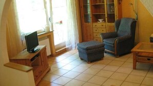 Uma sala de estar com televisão sobre um suporte de madeira, uma poltrona azul, uma mesa de centro com tampo de vidro e um armário de madeira com portas de vidro.