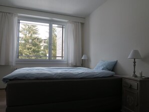 Een slaapkamer met een bed, twee nachtlampjes, een raam met gordijnen en een dressoir.
