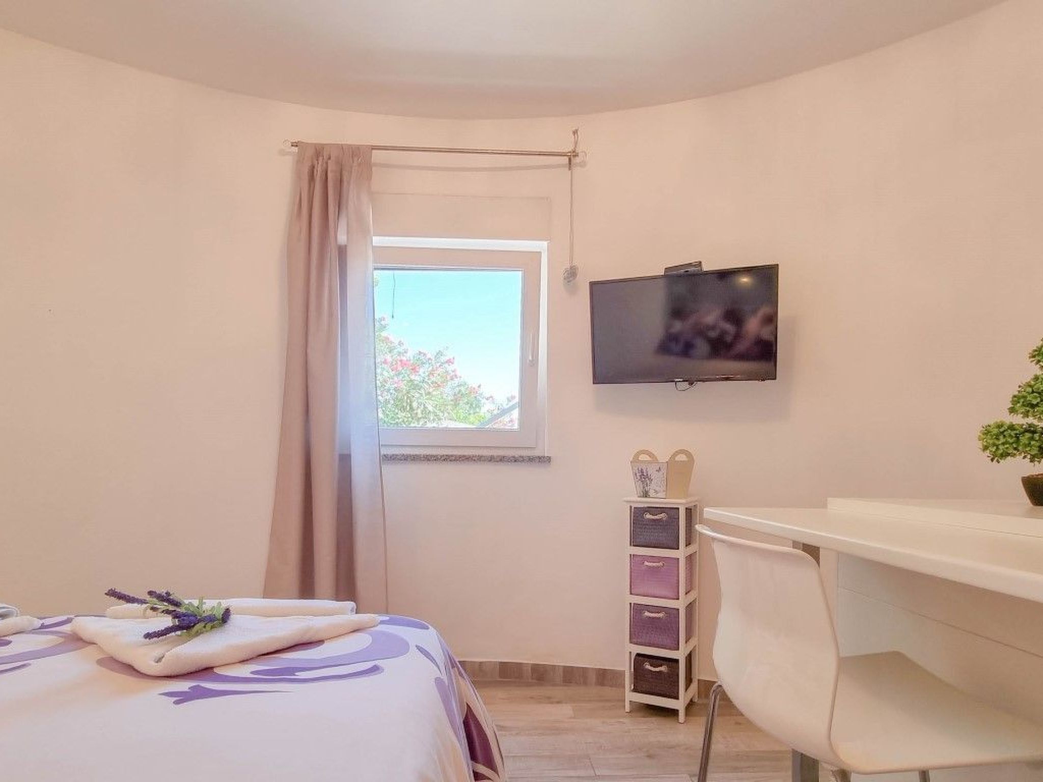 Un dormitori amb un llit, una taula de treball, una cadira, un televisor, una finestra amb cortines i una planta.