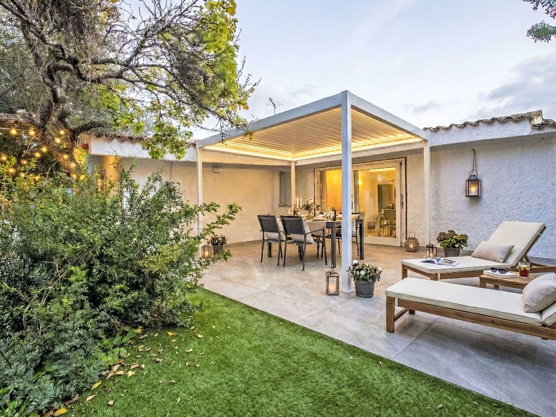 A covered patio with a dining table and chairs, a lounge area with a sofa and a chair, and a small garden with a tree and potted plants.