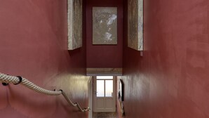 Um corredor estreito com uma corda à esquerda, uma escada de madeira que leva a uma porta no final e um quadro na parede.