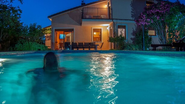 Ein Schwimmbecken mit einer Person im Wasser, umgeben von einem Haus mit Balkon und Gartenmöbeln.