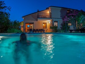 Ein Schwimmbecken mit einer Person im Wasser, umgeben von einem Haus mit Balkon und Gartenmöbeln.