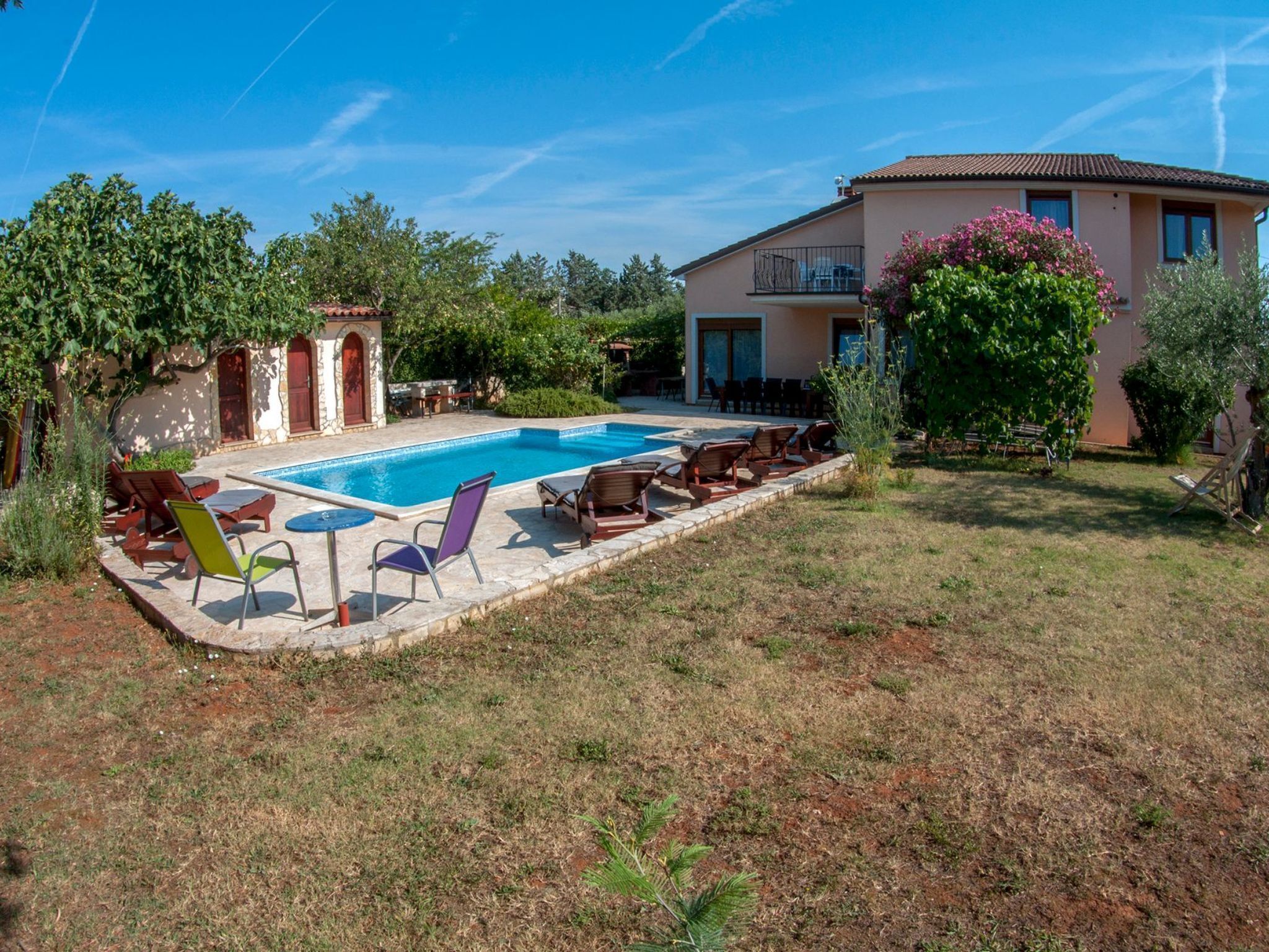 A swimming pool with lounge chairs and a patio area, surrounded by a garden and a building with a balcony.