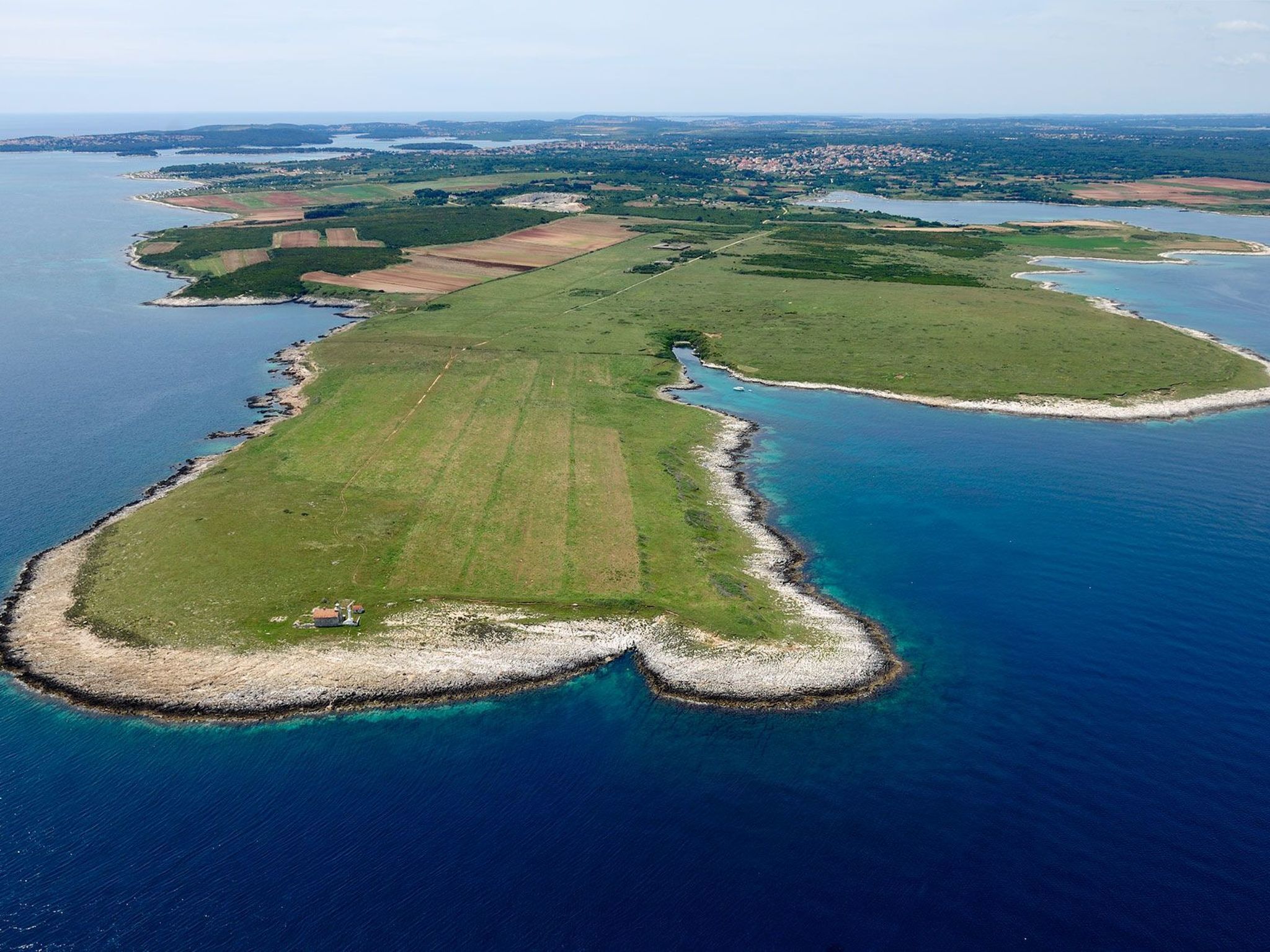 Vista aerea di una zona costiera con un ampio campo verde, un piccolo canale navigabile e una piccola isola con costa rocciosa.