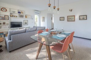 Un soggiorno moderno con tavolo da pranzo in vetro, divano grigio e poltrone rosse. Si nota una TV, mensole con oggetti decorativi e opere d'arte alle pareti.